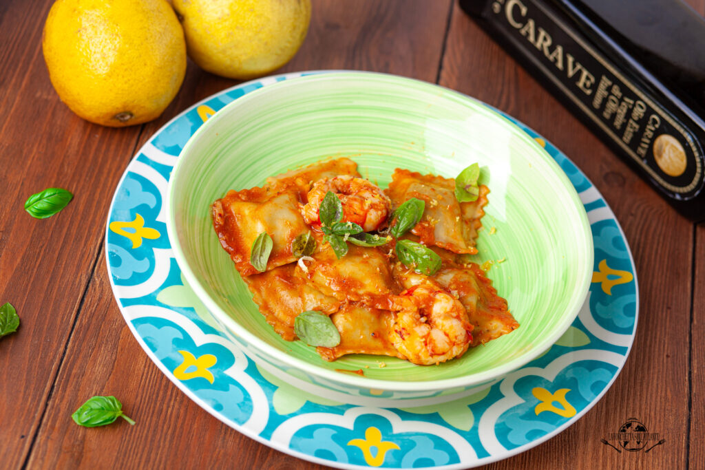 Ravioli fatti in casa al profumo di limone, conditi con bisque di gamberi e un filo di olio Carave, serviti su un piatto decorato con basilico.