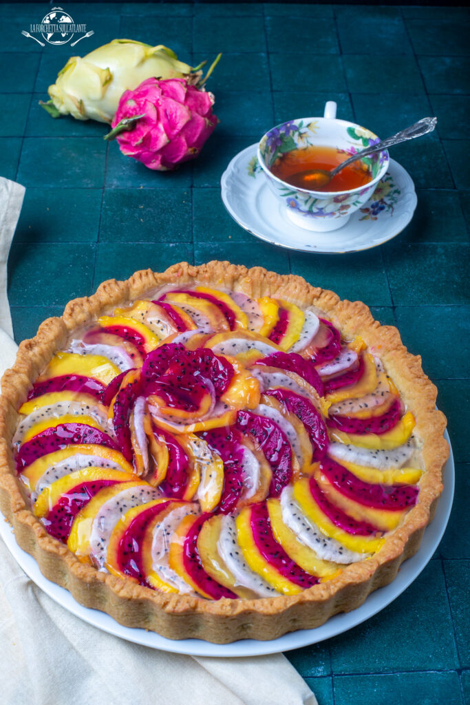 Crostata al mango e dragon fruit con crema pasticcera allo yuzu, decorata con fette di frutta tropicale bianca e magenta, lucidata con gelatina.

